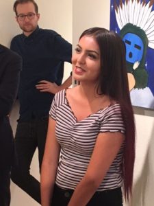 A teenaged girl with long black hair and dark eyes, speaking in front of a painting of a Hopi katsina with a tall blond man leaning against the wall behind her, listening