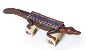 Painted wooden sculpture of an alligator resting on two block supports, decorated with red, blue, and geometric patterns.