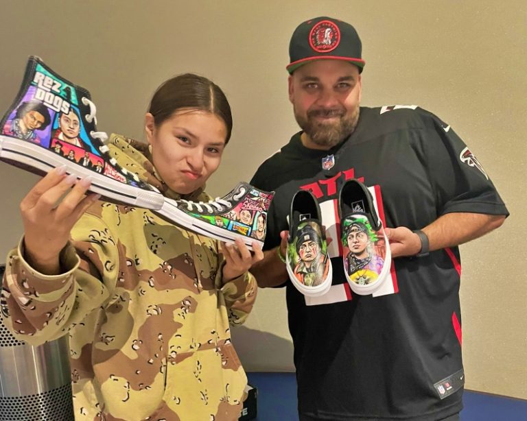 Paulina Alexis and Chris Thompson pose indoors holding custom hand-painted sneakers, including high-top Converse featuring Reservation Dogs characters and slip-ons with detailed portrait artwork.